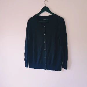 Svart kofta från Esmara med knappar strl M - En klassisk svart kofta från Esmara i storlek M. Koftan har rund halsringning, långa ärmar och stängs med knappar framtill. Perfekt att slänga över en t-shirt eller linne för en enkel och stilren look.  Helt skick dock noppror