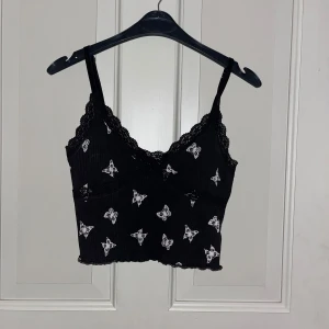 Svart linnetopp med fjärilar och spets - Supersöt svart linnetopp med vita fjärilar och spetskant vid urringningen. Toppen har smala axelband och croppad passform. Perfekt för dig som gillar detaljer och vill ha en trendig look med lite extra edge.