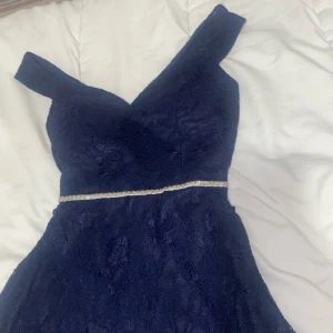 Marinblå spetsklänning med silverdetalj - Snygg marinblå klänning med spets och v-ringad offshoulder-överdel. Midjan markeras av ett glittrigt silverband som ger extra wow-känsla. Perfekt för bal eller fest. Klänningen har en figurnära passform upptill och utsvängd kjol.