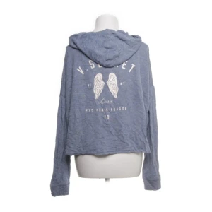 Blå hoodie från Victoria's Secret - Mysig blå hoodie från Victoria's Secret med dragkedja och huva. Tröjan har vita tryck med vingar och text på ryggen, samt två fickor framtill. Oversized passform och långa ärmar, perfekt för chill dagar.