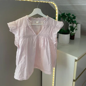 Ljusrosa blus från Gina Tricot - Supersöt ljusrosa blus från Gina Tricot med volangärmar och v-ringning. Blusen har fina rynkade detaljer upptill och en lös passform. Har fått en liten fläck i bak på ryggen! 