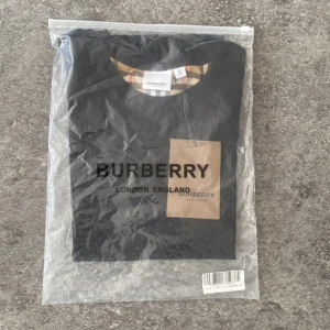 Burberry svart t-shirt med rutiga ärmar - Svart t-shirt från Burberry med klassiska rutiga ärmar i beige, rött och svart. T-shirten har rund halsringning och är tillverkad i mjuk bomull. Perfekt för dig som vill ha en stilren look med en touch av lyxig streetstyle.