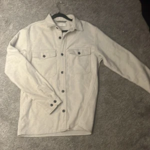 Beige overshirt från Jack & Jones M - Snygg beige overshirt från Jack & Jones Premium med klassisk krage, två bröstfickor med lock och svarta knappar. Tillverkad i mjukt material som känns lite som ull. Perfekt att slänga över en t-shirt för en chill look. Priset ej hugget i sten🙌
