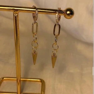 Guldiga statement örhängen med kedjor - Snygga guldiga örhängen med kedjedetaljer och spetsiga hängen. Örhängena har små gnistrande stenar på ringen och coola länkar som ger en edgy vibe. Perfekta för att lyfta vilken outfit som helst.