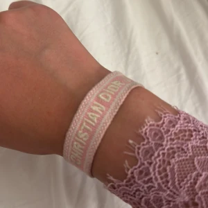 Christian Dior rosa armband - Superfint armband från Christian Dior i ljusrosa tyg med broderad text 'Christian Dior J'Adior' i vitt. Armbandet har justerbar knytning med tofsar i ändarna och en mjuk, casual vibe. Perfekt accessoar för att lyfta din outfit med en touch av lyx. Armbandet är inte äkta!!!!!