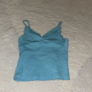 Blå ribbad linnetopp med spets - Supersöt blå linnetopp med smala axelband och v-ringning. Toppen har ribbad struktur, spetsdetaljer längs urringningen och en snygg rynkning framtill. Perfekt för varma dagar eller att styla lager på lager.