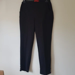 Svarta byxor från Noble Micha, strl 40 - Svarta byxor från Noble Micha i stretchigt material med smal passform. Byxorna har diskreta fickor och små knappar vid benslutet. Tillverkade i polyester, viskos och elastan för en bekväm känsla. Perfekta till en clean och stilren look.  Noppriga & färgsläpp på "knapparna" vid benen annars helt skick