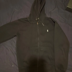 Svart hoodie från Polo Ralph Lauren - Svart hoodie från Polo Ralph Lauren med dragkedja och klassisk logga broderad på bröstet. Tröjan har huva med snörning och stora fickor framtill. Perfekt för en chill och stilren look.