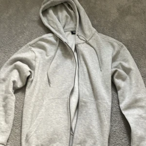 Ljusgrå hoodie med dragkedja 157 - Ljusgrå hoodie från 157 i storlek S. Tröjan har huva med snörning, dragkedja framtill och två fickor. Mjuk insida och klassisk passform, perfekt för chill dagar eller när du vill vara bekväm.