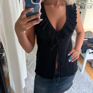 Svart plisserad blus från Zara - Supersnygg svart plisserad blus från Zara med djup v-ringning och bred volangkant runt halsen. Blusen är ärmlös och har knyt framtill 