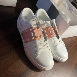 Valentino Garavani Rockstud sneakers - Snygga vita Valentino Garavani Rockstud sneakers med rosa band och ikoniska nitar över vristen. Skorna har rund tå, platt sula och är tillverkade i lyxigt skinn. Perfekt för dig som vill sticka ut med exklusiva detaljer och modern design. Säljaren som jag köpte dom ifrån säger att dom är äkta, jag köpte dom från Plick här! Aldrig använda🥰köpte för 2100kr
