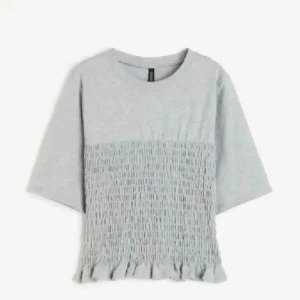 Ljusgrå topp med volangkant - Superfin ljusgrå topp med korta ärmar och smockad midja som ger en cool struktur. Volangkant nertill för extra detalj. Perfekt till jeans eller kjol för en chill och trendig look.