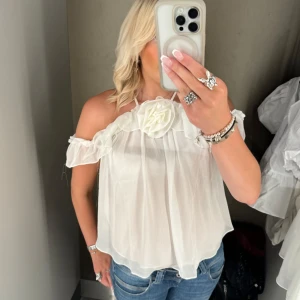 Vit blus med volang och blomdetalj - Superfin vit blus från Amisu med volangkant och stor tygblomma framtill. Ärmlös modell med tunna axelband och lätt transparent tyg som ger en luftig känsla. Perfekt för sommarens alla tillfällen när du vill känna dig extra söt. Tröjan är aldrig använd men lappen sitter dock inte kvar!