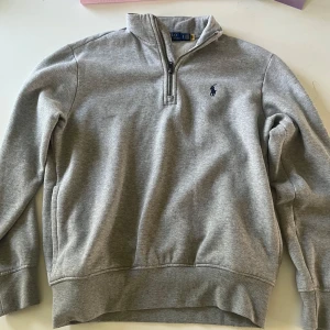 Grå half zip tröja från  Ralph Lauren - Snygg grå långärmad tröja från Polo Ralph Lauren med half zip och broderad logga på bröstet. Mjuk insida och klassisk passform, perfekt för lager-på-lager. Enkel att matcha med jeans eller chinos.