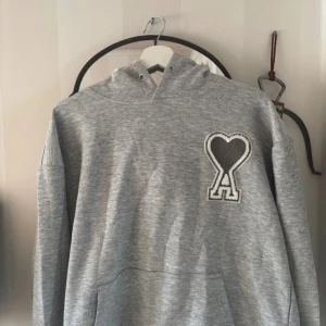 Grå hoodie från Ami med hjärtdetalj - Snygg grå hoodie från Ami Alexandre Mattiussi med stor svart hjärtdetalj och bokstaven A på bröstet. Klassisk huva och känguruficka framtill. Perfekt för dig som gillar stilrena och trendiga plagg. Jag tog bort snörena men de ba sätta tillbaka dem. Priset kan diskuteras 