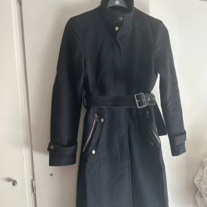 Svart kappa med bälte och dragkedjor - Snygg svart kappa med bälte i midjan och coola dragkedjor vid fickorna. Hög krage och långa ärmar med knäppning vid ärmslut. Perfekt för dig som vill ha en stilren och tidlös look under kallare dagar. Mycket fint skick.