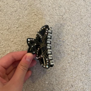 Svart hårklämma med strass och guld - Snygg svart hårklämma med glittriga strasstenar och tunna gulddetaljer. Perfekt accessoar för att lyfta din frisyr med lite extra bling. Klämman har vita tänder och ett elegant, stilrent utseende.