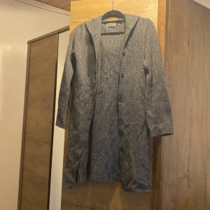 Grå stickad cardigan jacka KappAhl - Mysig grå stickad cardigan jacka från KappAhl med huva och knappar framtill. Lång modell med ribbade detaljer och diskret mönster. Perfekt för lager-på-lager och chill dagar. Passar dig som gillar en avslappnad och enkel stil.