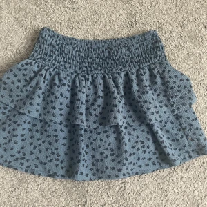 Blå volangkjol med blommigt mönster - Supersöt blå kortkjol med små svarta blommor och volanger. Den har även inbyggda shorts under volangerna. 