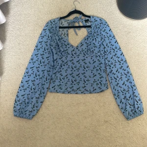 Blommig blå blus från Gina tricot - Supersöt ljusblå blus med svarta blommor från Gina Tricot . Toppen har v-ringning, långa ärmar med resår vid handlederna och snygg knytning i nacken som ger en öppen rygg. Perfekt för dig som gillar romantiska detaljer och croppad passform.