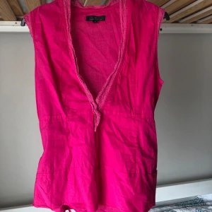 Ceriserosa ärmlös blus med knyt - Snygg ceriserosa ärmlös blus från ”DAY Birger et Mikkelsen” med djup v-ringning och spetskant. Ena sidan har knappar, vilket gör den lätt att komma i. Man kan knyta snöret på flera olika sätt och det går även att ta bort snöret helt.