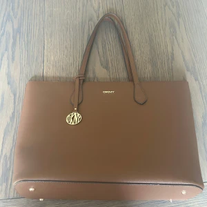 Brun handväska från DKNY i skinn - Snygg brun handväska från DKNY i stilren design med guldiga detaljer och logotyp. Väskan har två handtag, rymligt fack och dragkedja på insidan. Perfekt för dig som vill ha en snygg och praktiskt väska till skola eller jobb.