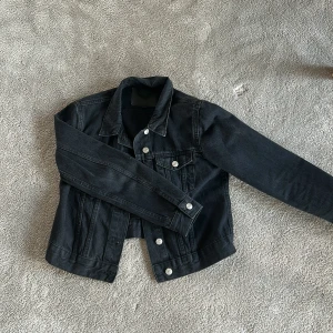 Svart jeansjacka från 157 XS - Svart jeansjacka från 157 i storlek XS. Klassisk modell med silvriga knappar framtill och på ärmslut, samt två bröstfickor med lock. Jackan har en rak passform och är tillverkad i slitstarkt denimtyg. Perfekt för dig som gillar en clean och tidlös look. Ganska kort i modellen. Ord. Pris 400 kr