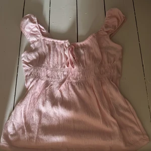 Ljusrosa blus med knyt och volang - Supersöt ljusrosa blus med knyt framtill och volangiga axelband. Perfekt för varma och soliga dagar när du vill känna dig extra gullig. Storlek Xs, fri frakt