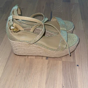 Glittriga guldiga sandaler Michael Kors - Snygga sandaler från Michael Kors med glittriga guldiga remmar och hög platåsula i repimitation. Skorna har öppen tå, justerbar rem runt vristen med guldfärgat spänne och plattform för extra höjd. Perfekta för att sticka ut med en touch av glamour. 