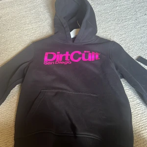 Svart hoodie Dirt Cült San Diego - Svart hoodie från Dirt Cült San Diego med stor magficka och huva. Tryck i metallicrosa på bröstet som verkligen poppar. Perfekt för dig som gillar streetwear och vill sticka ut lite extra. Skön och avslappnad passform.