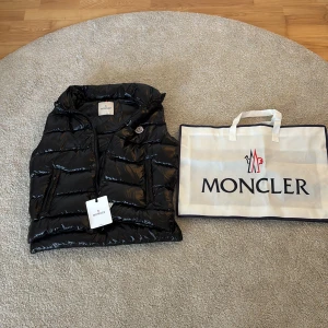 Svart dunväst från Moncler - Säljer en svart, glansig dunväst från Moncler med klassisk quiltad design och dragkedja framtill. Västen har två fickor med dragkedja och ikonisk Moncler-logga på bröstet. Perfekt för lager-på-lager och kommer med originalpåse.