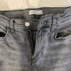 Grå jeans från Gina Tricot Perfect Jeans - Säljer ett par grå utsvängda jeans från Gina Tricot, modellen Perfect Jeans. De har en klassisk femficksdesign, dragkedja och knapp framtill samt råa, avklippta benslut för en cool vibe. Jeansen är i tvättad grå nyans och tillverkade i jeanstyg.
