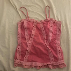 Rosa linnetopp med spets från Frime - Supersöt rosa linnetopp från Frime med tunna axelband och detaljerad spets längs kanterna och framtill. Toppen har en feminin volangkant nertill och är perfekt för en somrig look. Tillverkad i mjuk bomull som känns lätt och luftig.