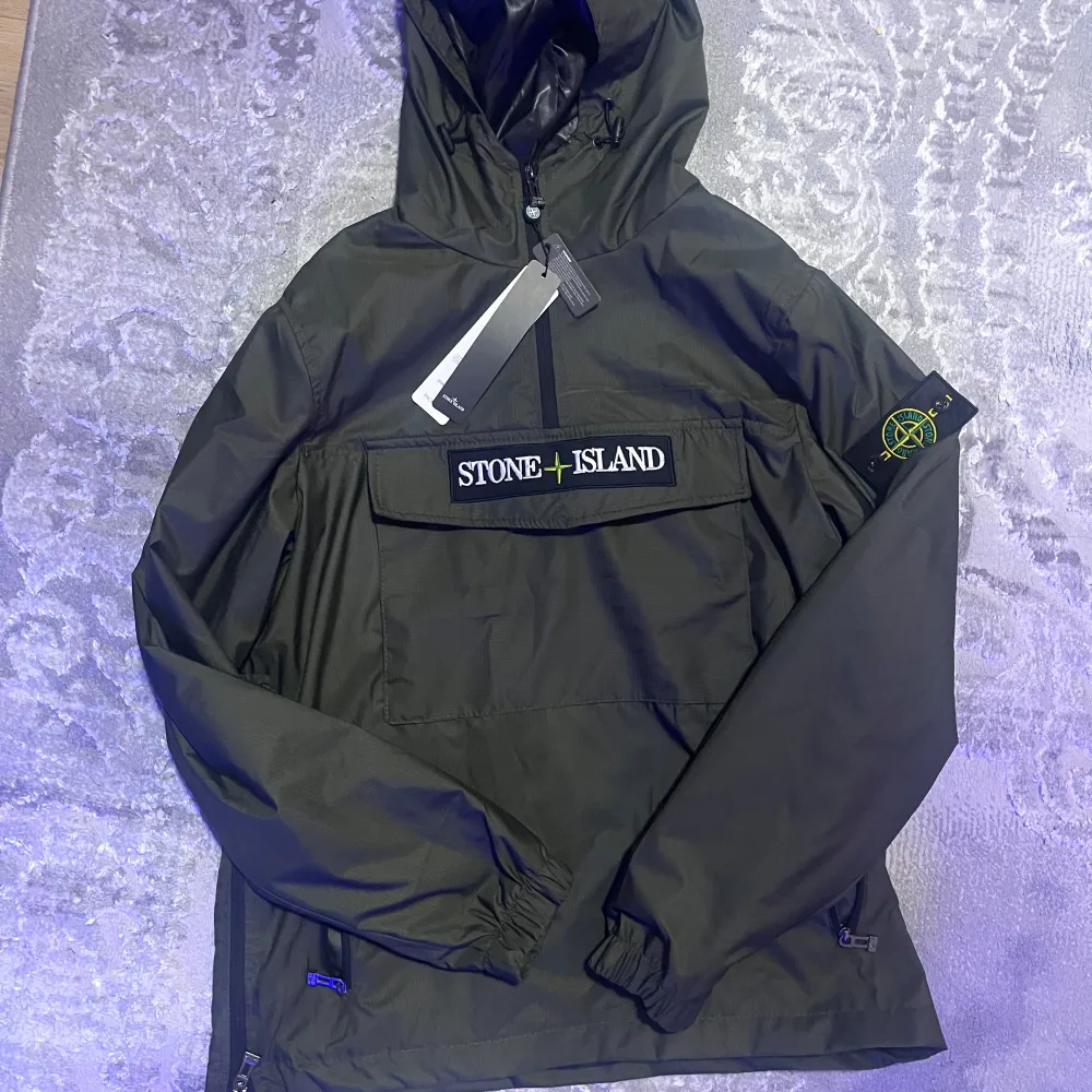 Snygg svart vindjacka från Stone Island med huva och dragkedja. Jackan har en stor ficka framtill med Stone Island-logga och en ikonisk patch på ärmen. Perfekt för blåsiga dagar.. Takit.