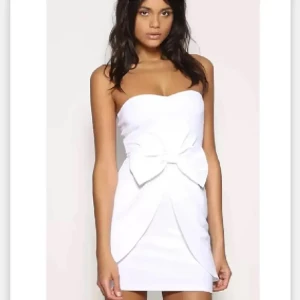 Vit axelbandslös klänning med rosett - Snygg vit axelbandslös klänning med en stor rosett framtill. Perfekt för en elegant look. Klänningen har en kort längd och en figurnära passform.
