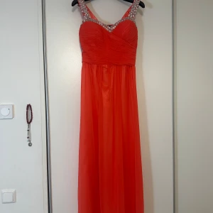 Röd långklänning med pärldetaljer - Snygg orange långklänning med pärldetaljer på axelbanden och runt halsringningen. Klänningen har en vacker drapering över bysten och en flytande kjol. Perfekt för en elegant kväll.