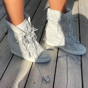 Ljusgrå boots med fransar - Snygga ljusgrå boots med fransar och snörning. Tillverkade i mocka med en rund tå och platt sula. Perfekta för en bohemisk stil.