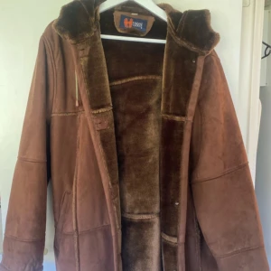 Brun mockajacka från Hudson - Säljer en snygg brun mockajacka från Hudson med huva och knappar framtill. Jackan har en mjuk insida och långa ärmar, perfekt för kyligare dagar. Den har en klassisk design med en bekväm passform. Tyvärr har en av knapparna ramlat av annars är det inget fel på den. 