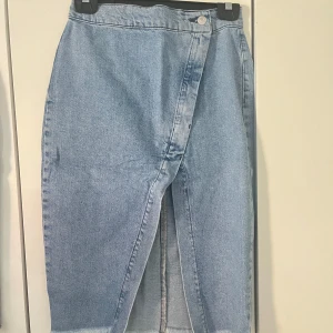 Blå jeanskjol med omlott - Snygg blå jeanskjol med omlott-design och knäppning fram. Kjolen har en slits framtill och två stora bakfickor. Perfekt för en avslappnad stil.