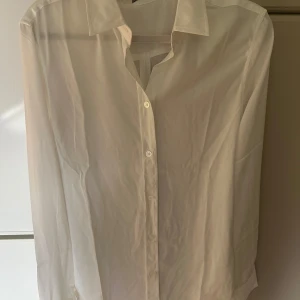 Vit långärmad skjorta - Säljer en klassisk vit långärmad skjorta med knappar framtill. Skjortan har en stilren design med krage och är perfekt för en elegant look. Materialet är lätt och bekvämt. Nytt och oanvänd 