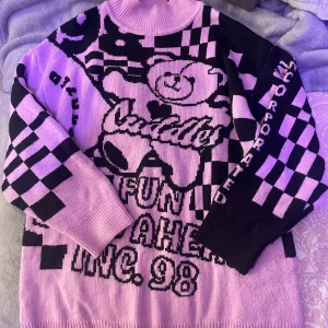 Alt Rosa svart tröja - Säljer en unik rosa och svart stickad tröja med ett coolt grafiskt mönster och textdetaljer. Tröjan har en hög krage och långa ärmar med rutigt mönster på sidorna. Perfekt för att sticka ut i mängden!