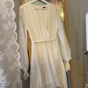 Vit prickig klänning från DM - Säljer en söt vit klänning från DM med prickigt mönster och omlott-design. Klänningen har långa ärmar med knytband vid ärmsluten och en v-ringad hals. Perfekt för sommarens fester!