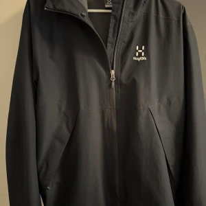 Svart vindjacka från Haglöfs - Säljer en stilren svart vindjacka från Haglöfs. Jackan har en praktisk dragkedja framtill och en justerbar huva. Perfekt för blåsiga dagar och utomhusaktiviteter. Den är designad för att ge bra skydd mot väder och vind.