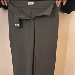 Grå byxor från Nelly - Snygga grå byxor från Nelly med låg midja och rak passform. Perfekta för en stilren look. Byxorna har en diskret dragkedja och knapp framtill.