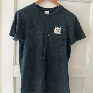 Mörkblå t-shirt med kattmotiv från Ripndip - Cool mörkblå t-shirt från Ripndip med ett unikt kattmotiv som gömmer sig i bröstfickan. Tillverkad i mjukt material för en skön passform. Den har ett litet hål vid fickan som har spruckit upp, se vänster sida av fickan på bild 2