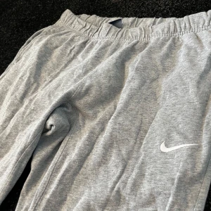 Grå mjukisbyxor från Nike - Säljer ett par grå mjukisbyxor från Nike med elastisk midja och en liten logga på benet. Perfekta för en avslappnad stil eller träning. Byxorna har en bekväm passform och är tillverkade i mjukt material.