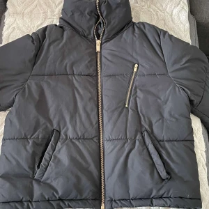 Svart pufferjacka med dragkedja - Säljer en svart pufferjacka från Ü med dragkedja framtill och en sned bröstficka med dragkedja. Jackan har en hög krage och två sidofickor. Perfekt för kyliga dagar!