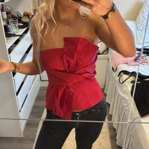 Röd axelbandslös topp med rosett - Säljer en snygg röd axelbandslös topp med en stor rosett framtill. Toppen har en elegant omlott-design och är perfekt för en kväll ute. Den är figurnära och ger en stilren look. Köpte här på plick men tyvärr inte stor på mig🥰