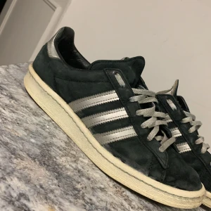 Adidas sneakers i svart mocka och silvriga ränder - Snygga Adidas sneakers i svart  mocka med klassiska tre ränder i silver på sidan. Skorna har en vit sula och snörning framtill. Perfekta för en avslappnad stil.  Tveka inte vid frågor 👌