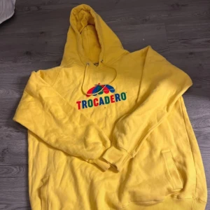 Gul hoodie från Trocadero - Säljer en gul hoodie från Trocadero med färgglatt tryck på framsidan. Hoodien har en klassisk design med känguruficka och justerbar huva med snören. Perfekt för en avslappnad stil. 🌟